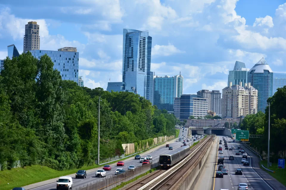 MARTA train tracks run in the middle of a six lane highway with an overpass and the Buckhead city skyline of skyscrapers in the background.